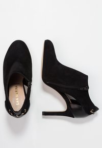 Bottines en daim noir avec un bout pointu, un haut talon aiguille et une fermeture zippée sur le côté. Présente des éléments en métal doré.