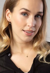 Collier avec pendentif en forme d'étoile dorée sur une chaîne délicate, présenté sur une chemise noire, associé à de petites boucles d'oreilles en or avec des perles.