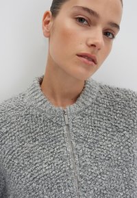 Texturierter grauer Pullover mit hohem Kragen und durchgehendem Reißverschluss. Weiches Material mit einem abwechslungsreichen Strickmuster, das einen leichten Glanz vom Reißverschluss aufweist.