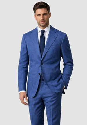 Uomo che indossa un abito blu su misura, camicia bianca e cravatta blu scuro, in piedi con una mano in tasca su uno sfondo chiaro uniforme.