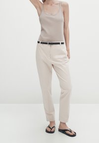 Beige tanktop, crèmekleurige broek met riemlussen en een zwarte riem, gecombineerd met zwarte slippers, tegen eenminimalistische achtergrond.