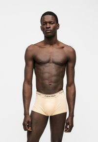 Νέος μυώδης μαύρος άνδρας που στέκεται μπροστά φοράει ανοιχτό μπεζ μποξεράκια Calvin Klein σε απλό λευκό φόντο.