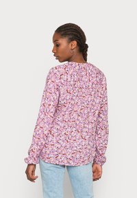 Blusa a maniche lunghe in tessuto viola con stampa floreale; presenta un dettaglio arricciato sulla schiena e polsini elasticizzati, abbinata a jeans azzurri chiari.