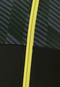 Gros plan sur un tissu comportant des sections vert foncé et noires avec une fermeture éclair jaune. Une texture à motif en lignes grises crée un intérêt visuel.