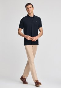 Marinblå polo skjorta, kortärmad, med tvåknappsknäppning, logotyp på bröstet, matchad med ljusbeiga byxor och bruna skor. Modell står med händerna hopknäppta.