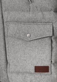 Hall villa kangas tekstuurse välimusega, millel on eesmine klapiga tasku, hõbedane nööp ja pruun nahast silt.