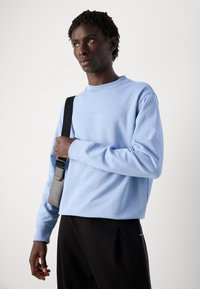 Joven con cabello corto y rizado, vistiendo sudadera azul clara y pantalones negros, sosteniendo la correa de un bolso cruzado gris y negro.