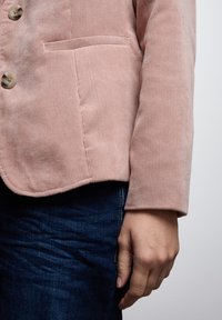 Růžový kordový blazer s knoflíkovým zapínáním, strukturovanou textilní látkou a náprsní kapsou, zkombinovaný s tmavě modrými džíny.