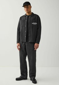 Schwarze Button-Up-Jacke mit zwei Fronttaschen, mit einem bedruckten Logo. Kombiniert mit dunklen Jeans und schwarzen Schuhen mit dicker Sohle.