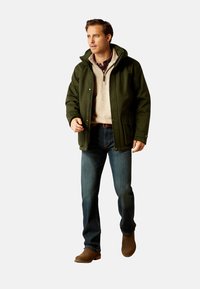 Grøn uldblandet jakke med hætte, beige sweater, blå jeans og brune støvler. Jakken har trykknaplukninger og sidelommer.