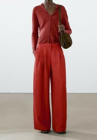 Osoba w czerwonym swetrze zapinanym na guziki i szerokich, czerwonych spodniach, trzymająca brązową torebkę na ramię, stojąca na jasnej podłodze z białym tłem.