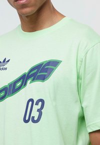 Šviesiai žalia medvilninė marškinėliai su dideliu mėlynos ir žalios spalvos "adidas" logotipu bei skaičiumi "03" ryškiomis mėlynomis skaitmenimis, apvalaus kaklo dizainas.