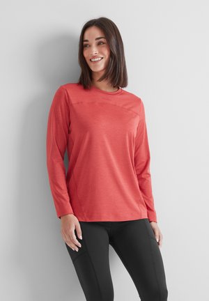 Woman smiling, wearing a long-sleeve coral top and black fitted pants, standing against a plain light gray wall.