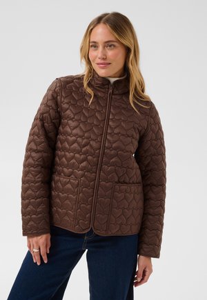Femme portant une veste matelassée marron avec des motifs en forme de cœur, debout avec une main posée à ses côtés et souriant légèrement.
