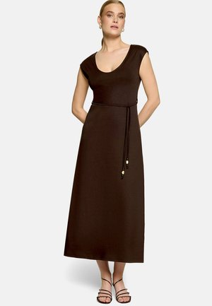 Femme portant une robe maxi marron foncé sans manches avec un décolleté rond et une fine ceinture à détails de perles, associée à des sandales noires à lanières.