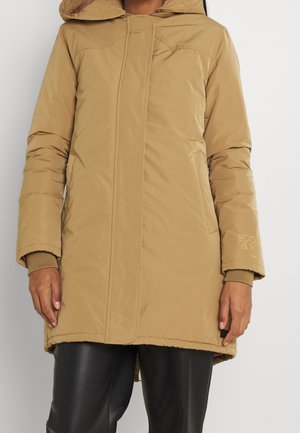 Femme portant un manteau matelassé à capuche couleur beige, longueur genou, par-dessus un pantalon noir, debout avec les bras détendus le long du corps sur un fond uni.