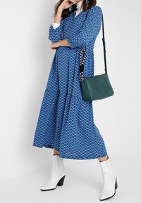 Blå mönstrad klänning med långa ärmar och flerlagrad kjol, tillsammans med en grön handväska. Vita ankelskor med blockklackar fulländar outfiten.