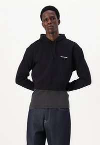 Svart kort hoodie med snøring og hette, med en hvit logo. Kombinert med mørke, strukturerte grå og svarte bukser, som fremhever kontrasten.