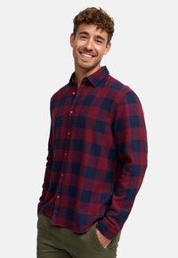 Camisa de franela a cuadros rojos y marinos con collar de botón, mangas largas y un bolsillo en el pecho. Se lleva con pantalones color verde oliva.