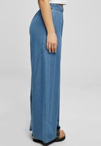 Frau trägt hoch taillierte, weit geschnittene blaue Jeans mit elastischem Taillenbund und schwarzen Plateausandalen vor grauem Hintergrund.