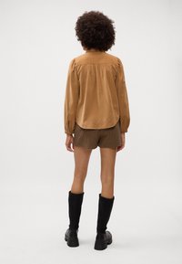 Camicetta a maniche lunghe in tessuto morbido marrone con colletto, dettaglio arricciato sul retro, abbinata a pantaloni corti a fantasia a quadri e stivali neri fino al ginocchio.