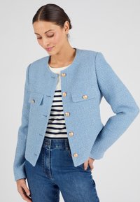 Lichtblauwe textuurjacket met een ronde hals, gouden knopen en twee voorzakken, gecombineerd met een gestreept shirt en een donkere spijkerbroek.