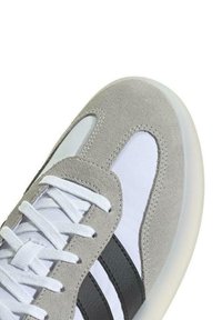 Zapatilla de ante gris con acentos blancos, que presenta tres franjas negras, una punta redondeada y una suela de goma texturizada. Los cordones son blancos.