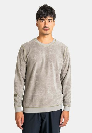 Mann mit kurzen dunklen Haaren, der einen hellgrauen, langärmeligen, strukturierten Sweatshirt und schwarze Hose trägt, steht vor einem einfarbigen Hintergrund.