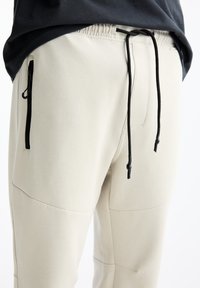 Helle beige Jogginghosen aus weichem Material mit schwarzen Reißverschlusstaschen, einem elastischen Bund und einer Kordel für eine verstellbare Passform.
