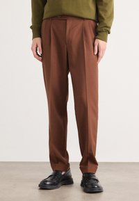 Braune maßgeschneiderte Hosen mit glatter Textur, mit einem gefältelten Design und einem klassischen Schnitt. Kombiniert mit schwarzen Slippern.
