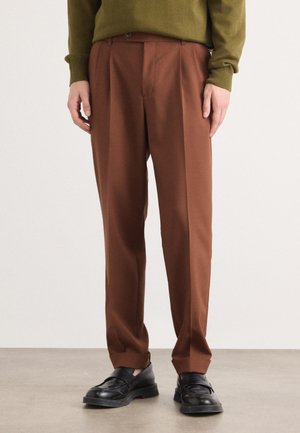 Brown tailored trousers with a smooth texture, featuring a pleated design and a classic fit. Paired with black loafers.
