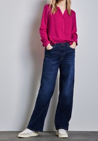 Fuchsia skjorta med knappar och långa ärmar, matchad med mörkblå vida jeans och vita sneakers med texturerade accenter.