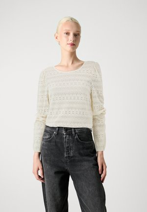 Lace long-sleeve top in off-white, featuring a round neckline and intricate floral patterns, paired with high-waisted black jeans.