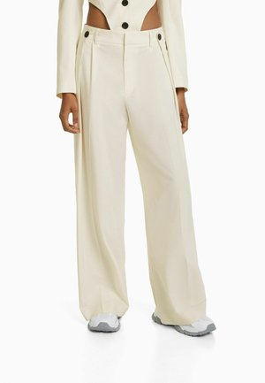 Pantalons à jambe large dans une couleur crème claire, dotés d'une taille haute, de plis et de boutons décoratifs. Le tissu semble lisse avec un design structuré.