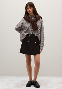 Camisa grande de cuadros blanco y negro con bufanda de punto marrón, combinada con una minifalda de tweed negra con botones dorados y mocasines negros.