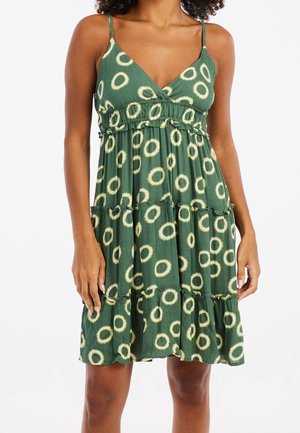 Kobieta w zielonej sukience bez rękawów z białym okrągłym wzorem i falbanami na dole, stoi z rękami swobodnie opuszczonymi.