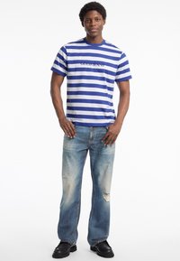 Mėlynos ir baltos juostelės marškinėliai su "GUESS JEANS" užrašu, derinami su šviesiai mėlynomis suplyšusiomis džinsais ir juodais batais. Lygus audinys, laisvas kirpimas.