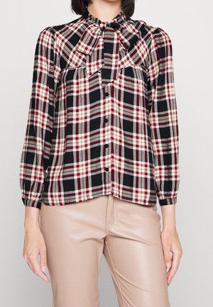 Kobieta ubrana w koszulę w kratę z długim rękawem i guzikami, z dużą kokardą przy szyi, w połączeniu z beżowymi spodniami.