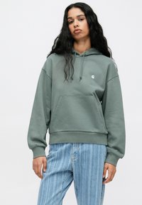 Een capuchontrui in gedempt groen, gemaakt van zacht materiaal. Beschikt over een voorkantzak en geribbelde manchetten. Gepaard met lichtblauwe gestreepte jeans.