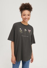 Giovane donna sorridente che indossa una T-shirt nera oversize con tre fiori e la scritta "YOU GROW, GIRL!" abbinata a jeans blu.