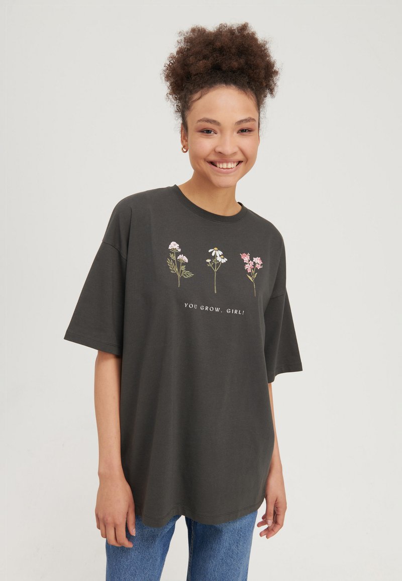 Giovane donna sorridente che indossa una T-shirt nera oversize con tre fiori e la scritta "YOU GROW, GIRL!" abbinata a jeans blu.