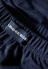Zbliżenie granatowego elastycznego pasa z czarną metką przyszytą od wewnętrznej strony, na której widnieje biały napis "LINDBERGH".