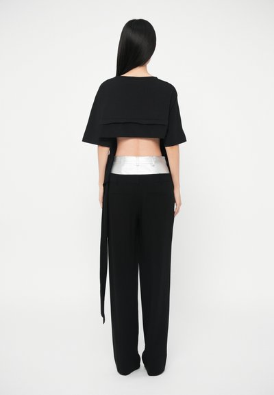 Naine, kellel on pikad mustad juuksed, seisab seljaga, seljas must lühike topp ja püksid, millel on hõbedane vöödetail, valge tausta ees.