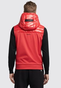 Rød puffervest med høj krave, glat tekstur og sorte detaljer. Har et logo på ryggen og ensfarvede sorte ærmer.