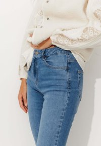 Jean en denim bleu taille haute avec une silhouette ajustée, présentant une couture discrète le long des poches et une fermeture à bouton en métal classique.