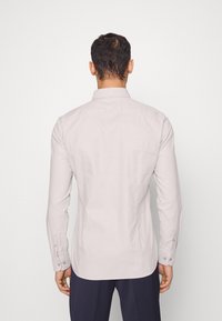 Camicia grigia chiara a maniche lunghe con colletto a bottoni e polsini; texture morbida e liscia; design aderente, vista posteriore mostrata su sfondo bianco.