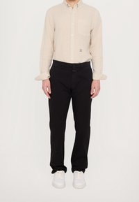 Uomo con camicia beige a maniche lunghe e bottoni, pantaloni neri e sneakers bianche che si trova davanti a uno sfondo bianco semplice.