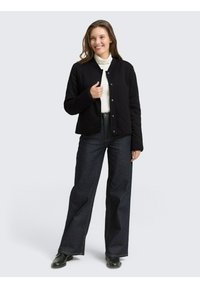 Zwarte gewatteerde jas met drukknopen, gedragen over een witte coltrui, gecombineerd met donkere jeans met wijde pijpen en zwarte schoenen. Neutrale achtergrond.