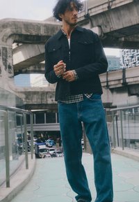 Črna jakna z gumbi, oblečena čez karirasto flanelasto srajco, v kombinaciji z modrimi jeans hlačami. Urbana postavitev z betonskimi strukturami v ozadju.