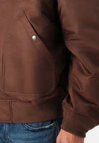 Casaco bomber castanho com um acabamento suave e brilhante, apresentando um bolso com botão e punhos canelados. Usado sobre jeans de ganga.